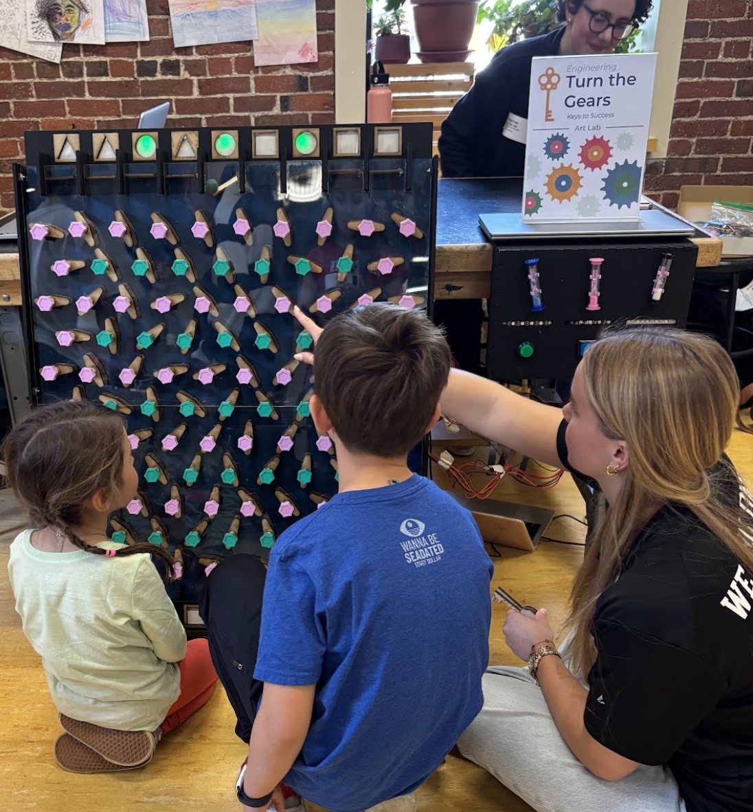 Two children playing Turn the Gears at the museum exhibit while a team member helps